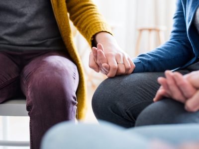 Midsection of men and women sitting in a circle during group therapy, supporting each other and holding hands.